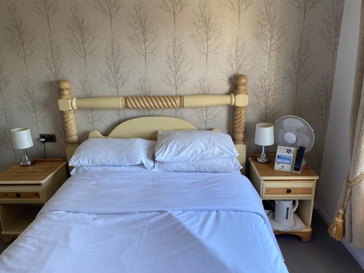 Una cama bien hecha con sábanas blancas, dos mesitas de noche con lámparas, un ventilador y un control remoto sobre una de las mesitas.