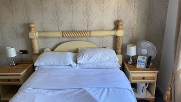 Una cama bien hecha con sábanas blancas, dos mesitas de noche con lámparas, un ventilador y un control remoto sobre una de las mesitas.