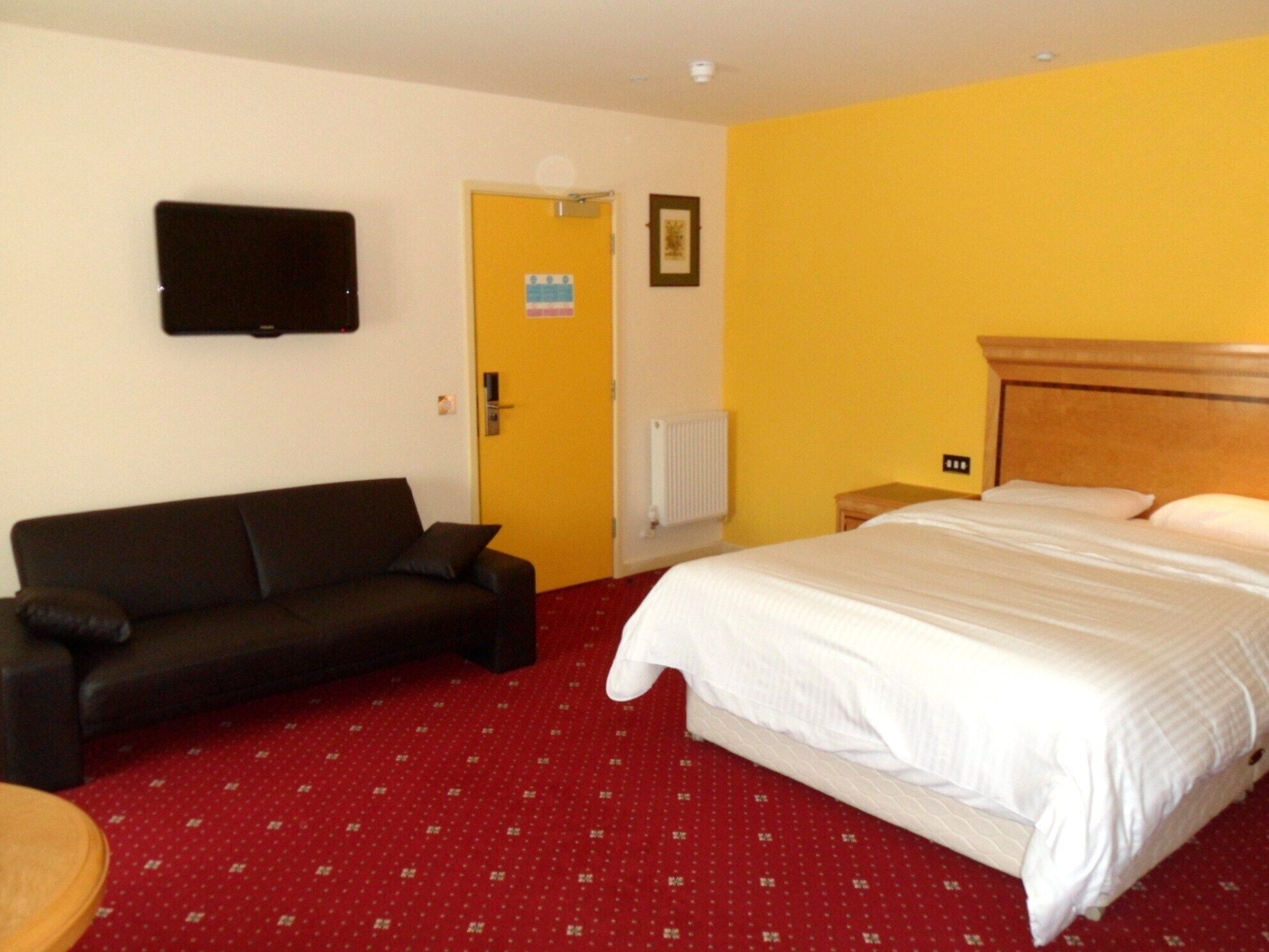 A hotel room with a bed, a sofa, a television mounted on the wall, and a door with a sign.