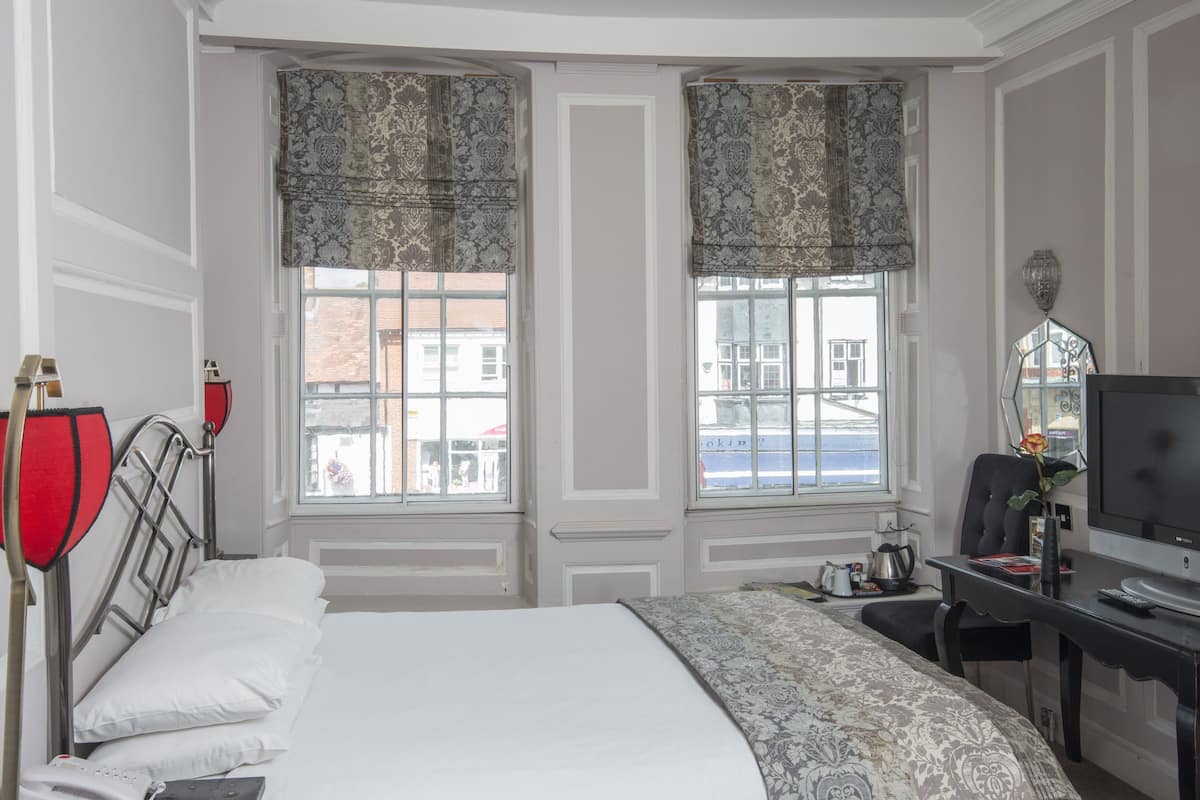 Habitación de hotel con una cama grande, dos ventanas con cortinas estampadas, un escritorio con televisión y vistas a edificios.