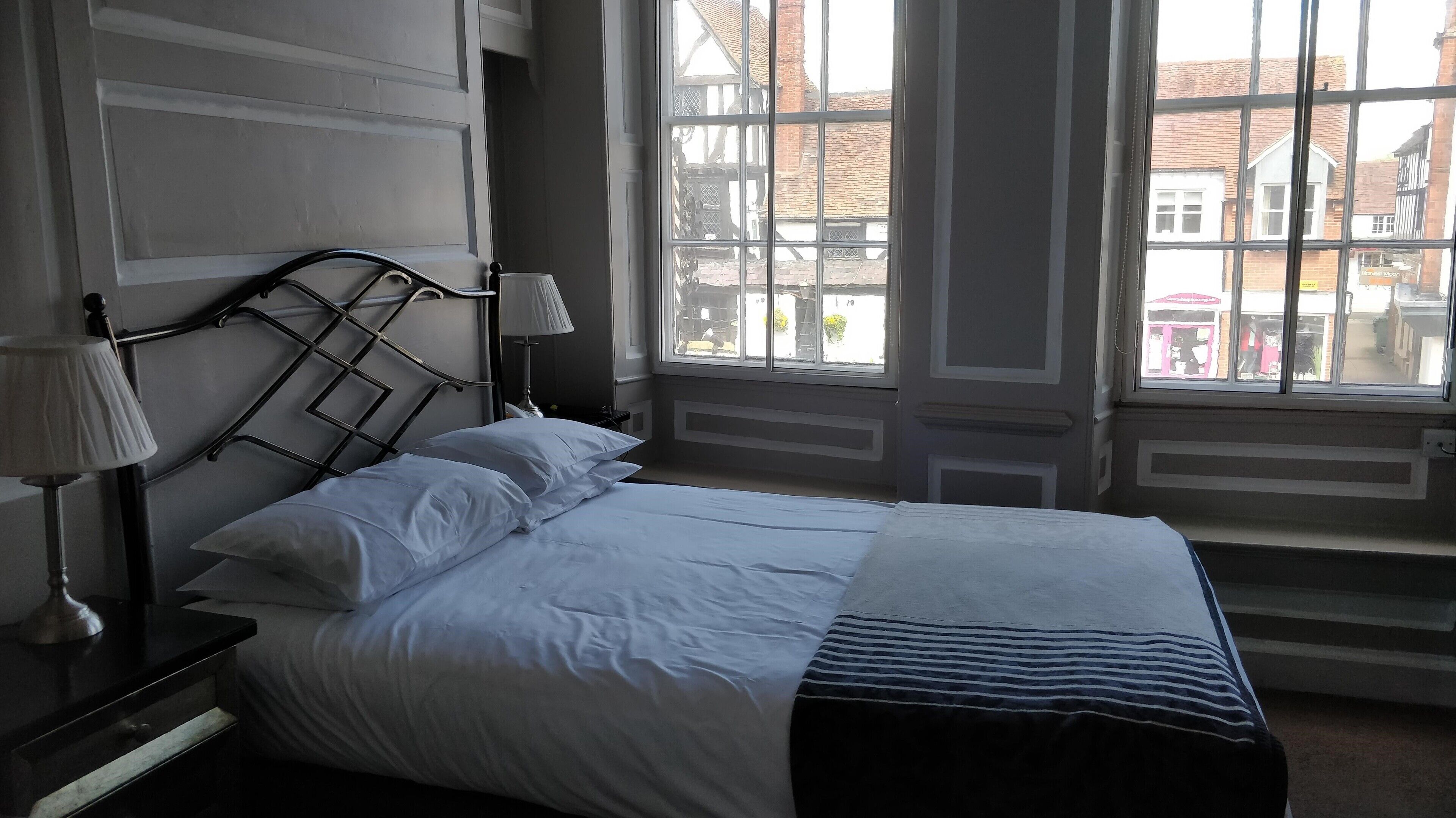 A neatly made bed with white linens and a striped blanket, a bedside lamp, and a view of the street through large windows.