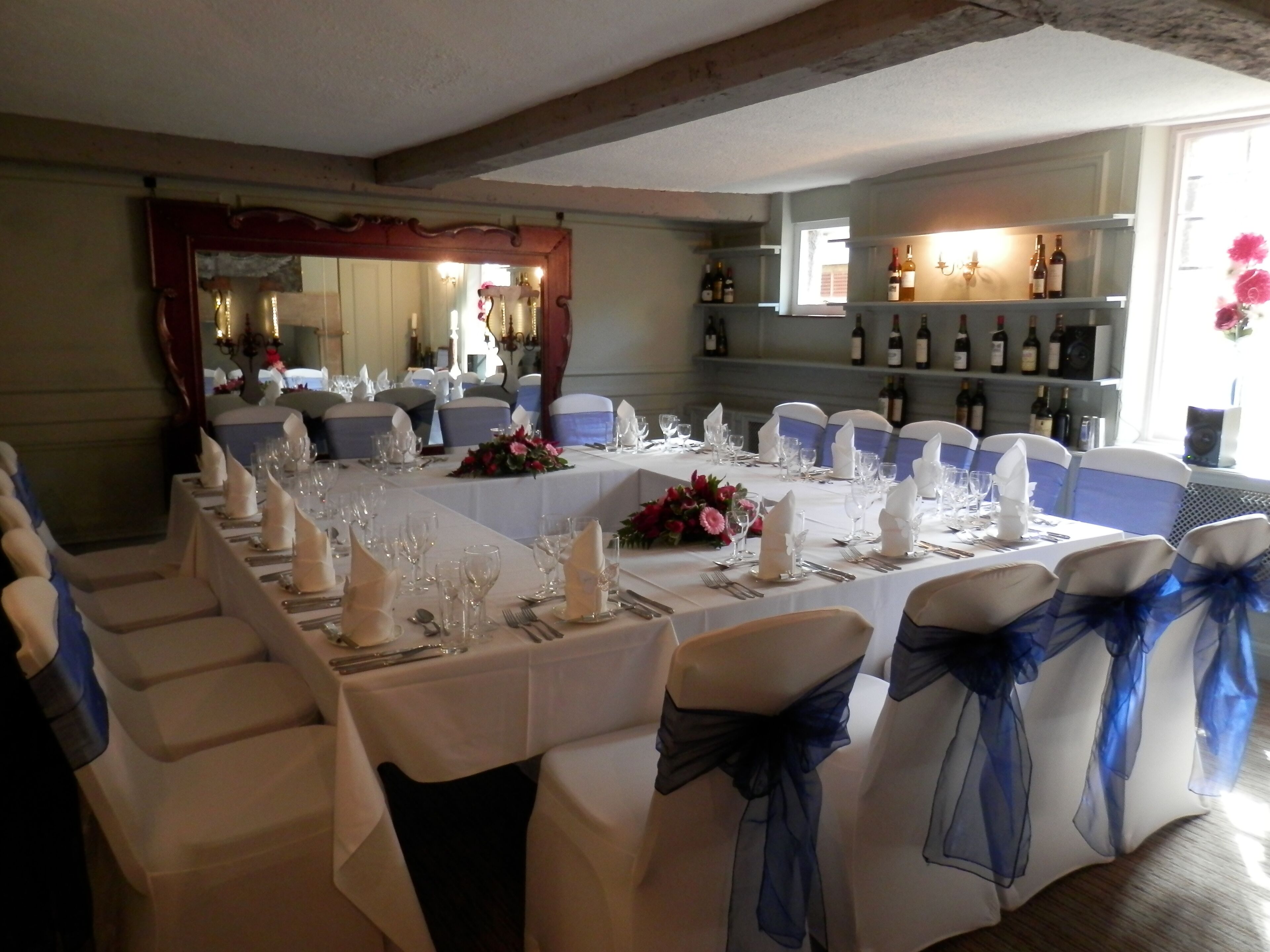 A long dining table set for a formal event, with white tablecloths, blue sashes on chairs, and wine bottles on a shelf in the background.
