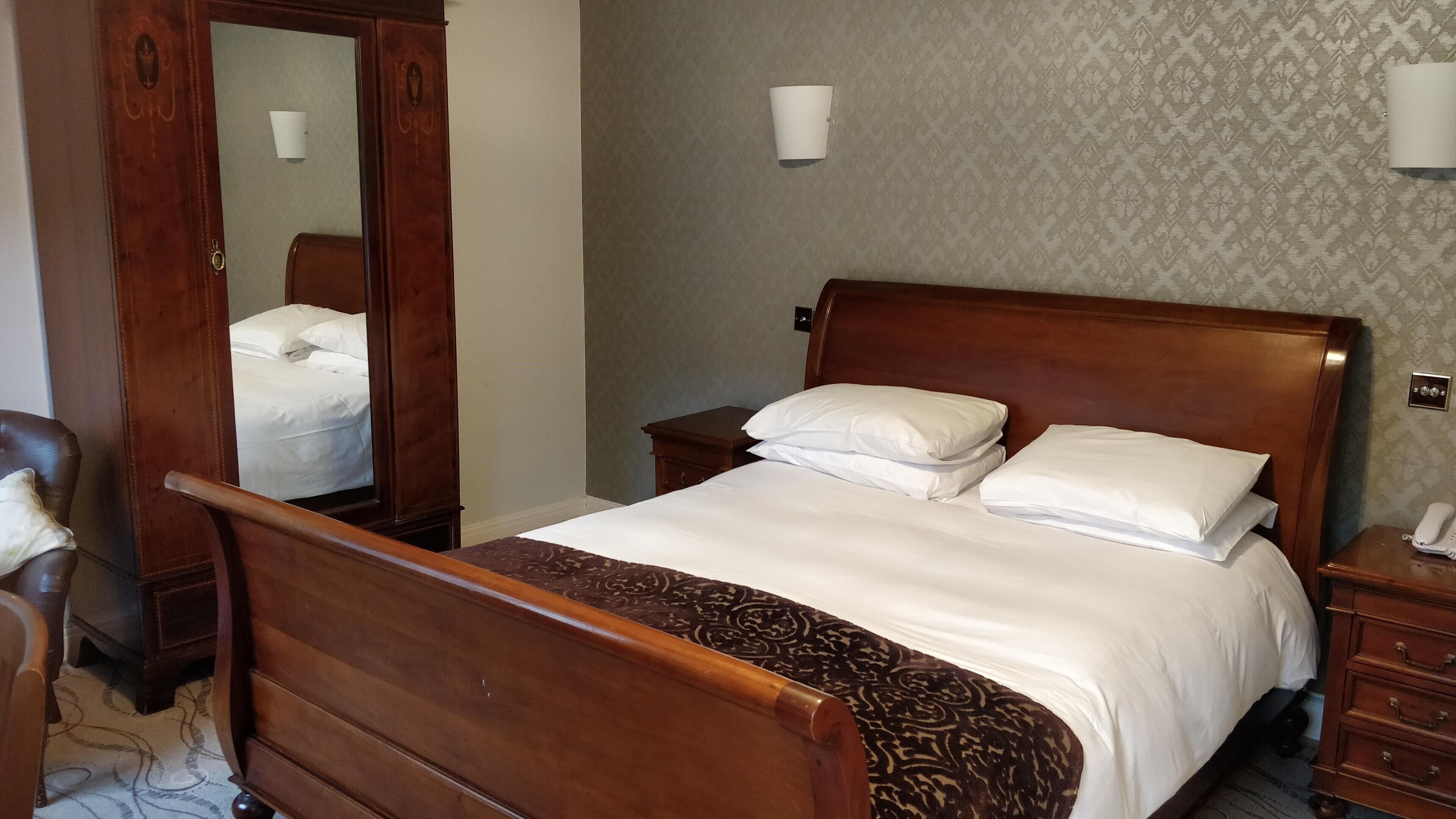 A wooden bed with white bedding, a nightstand with a phone, and a mirror on the wall.