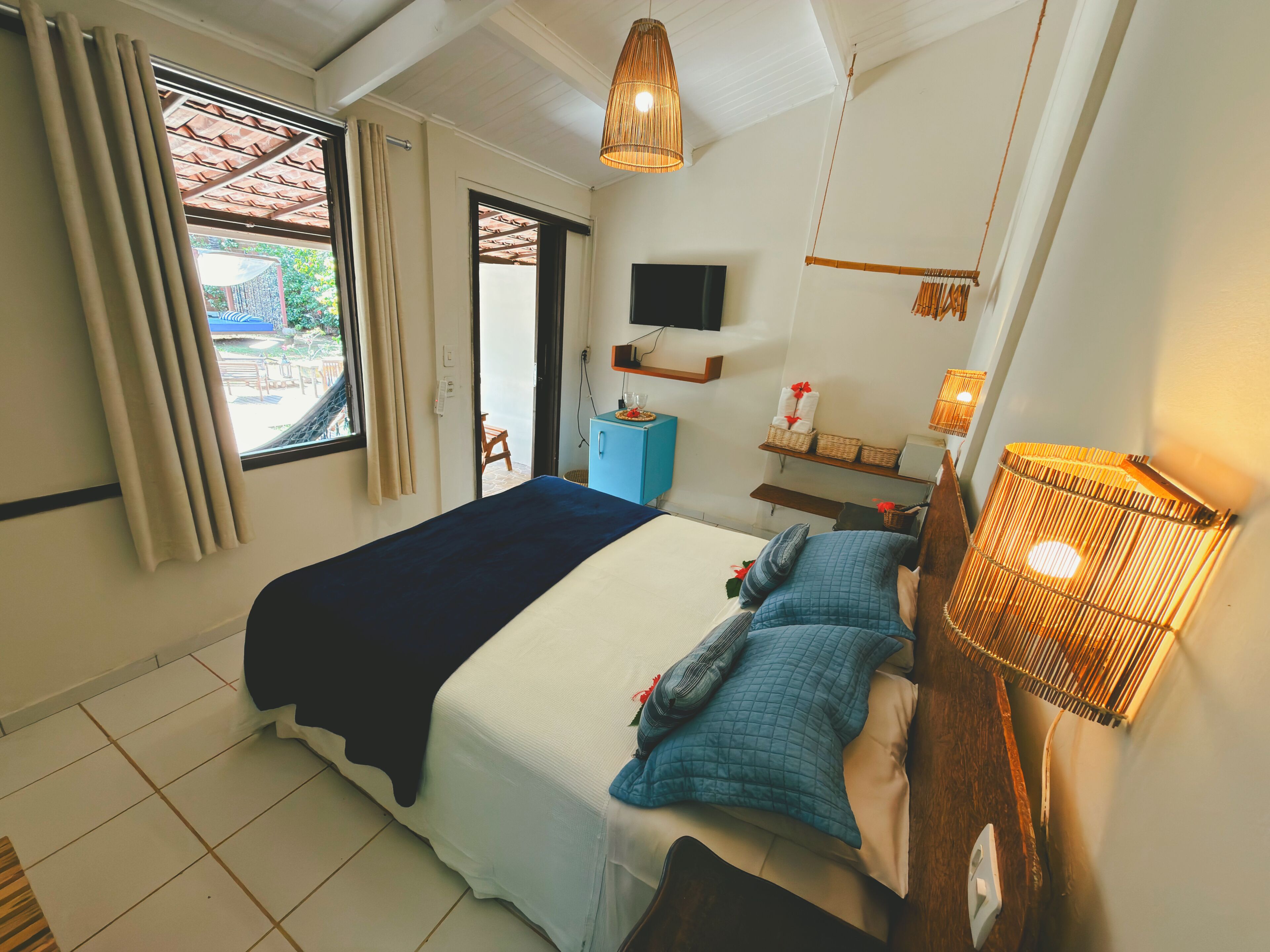 Quarto com cama, televisão, mesa de cabeceira, abajur e janela com cortinas.