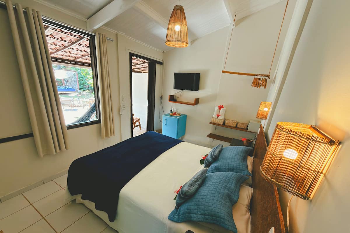 Quarto com cama, televisão, mesa de cabeceira, abajur e janela com cortinas.