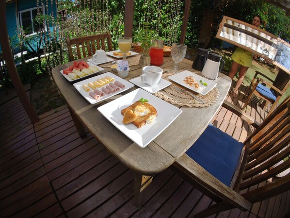 Une terrasse en bois avec une table mise pour le petit-déjeuner, comprenant des assiettes de nourriture, un verre de jus de fruits et une tasse.