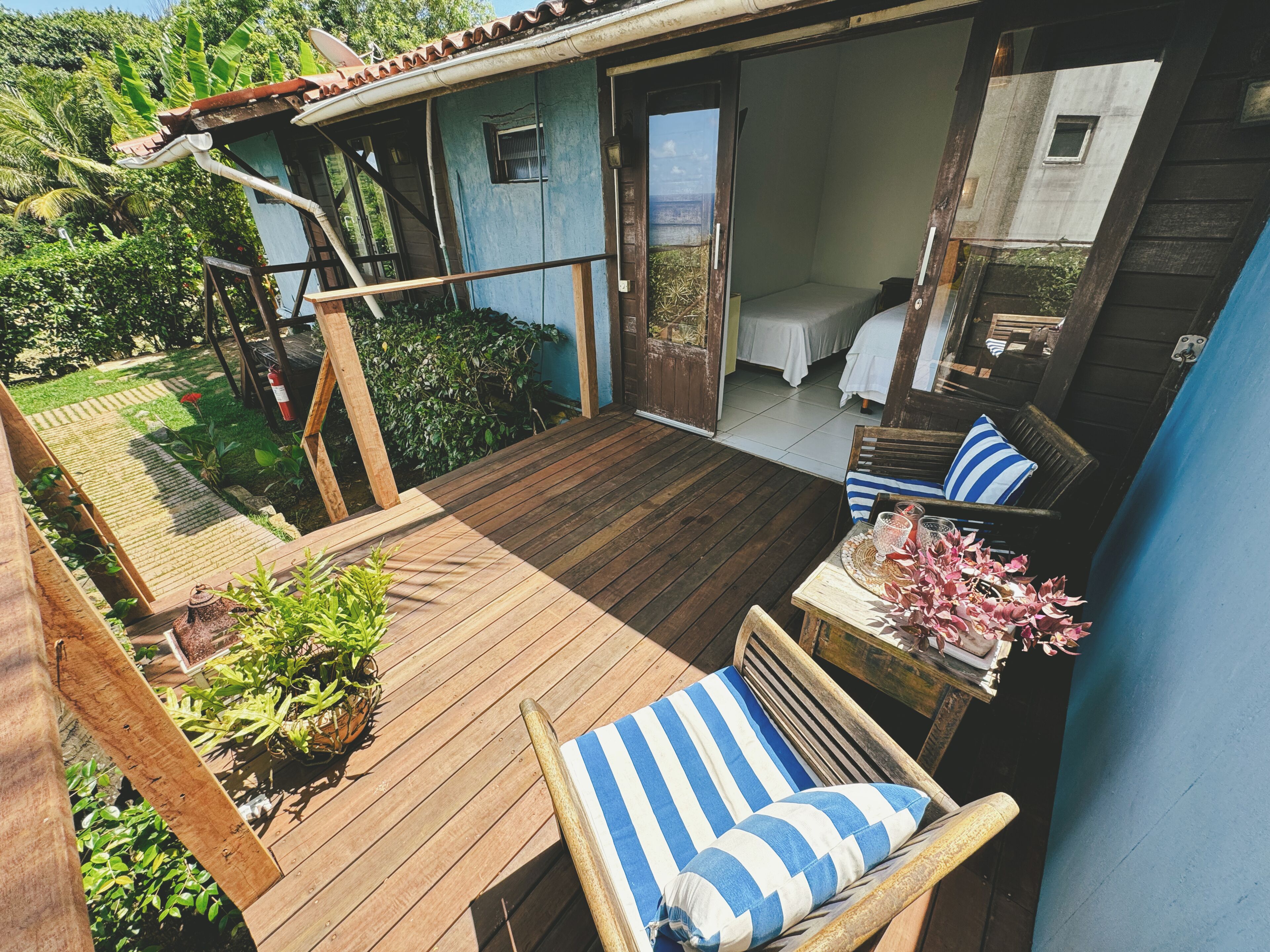 Una terraza de madera con una silla y una mesa, un pequeño jardín con plantas, y un edificio de fachada azul con una puerta que da a una habitación con cama.