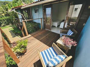 Eine Holzterrasse mit Stuhl und Tisch, ein kleiner Gartenbereich mit Pflanzen und ein Gebäude mit blauer Fassade sowie einer Tür zu einem Zimmer mit einem Bett.