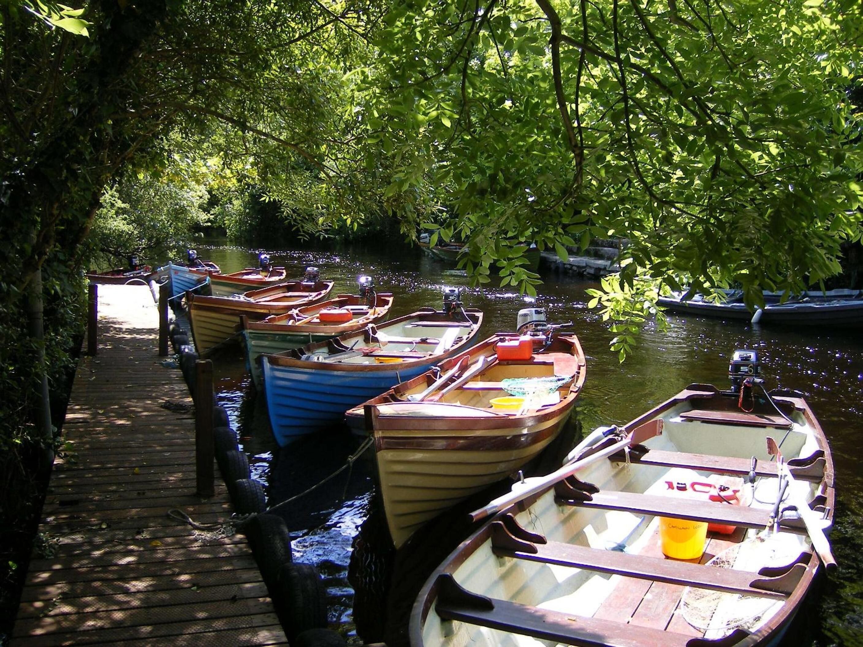 Une jetée en bois, où sont amarrés plusieurs petits bateaux, le long d’une rivière paisible, le tout entouré d’une végétation luxuriante.