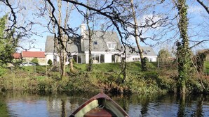 A narrow waterway with a rowboat in the foreground, a house with a red roof and stone walls in the background, and a grassy area with trees and shrubs. - Camillaun Lodge (Oughterard)