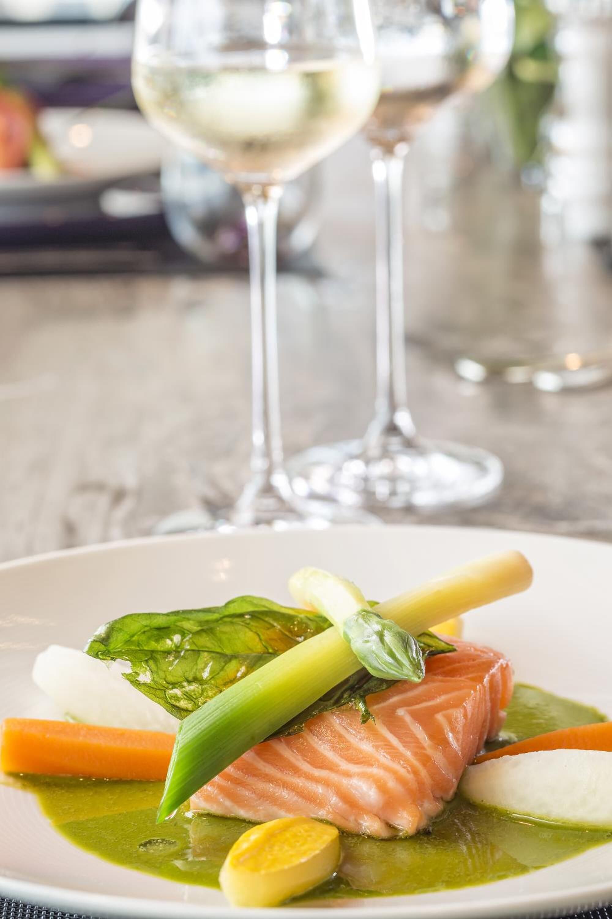 A plate of food with salmon, vegetables, and a green sauce, with two glasses of white wine in the background.