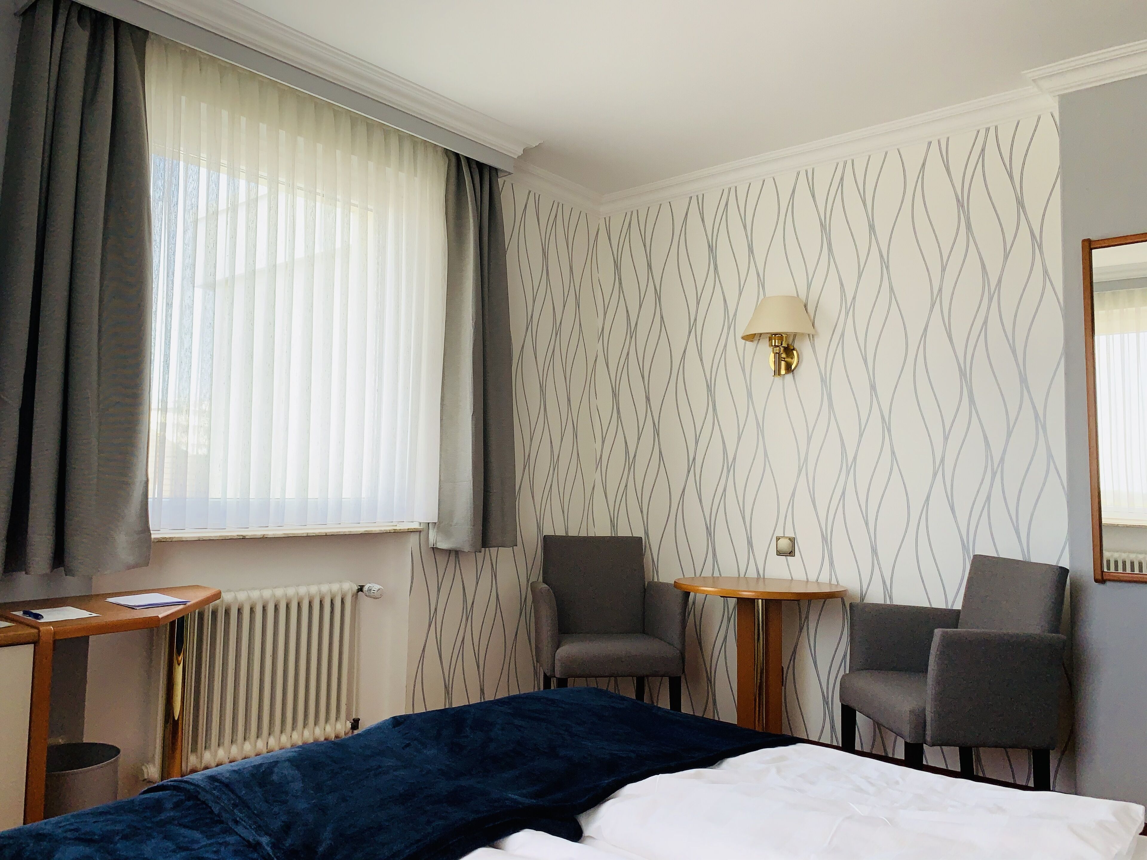 A hotel room with a bed, two armchairs, a small table, a lamp, and a radiator.