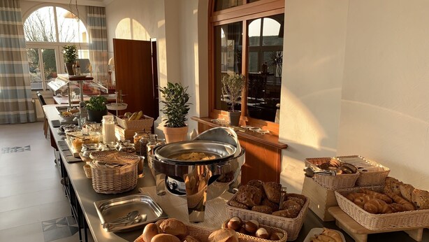 A buffet setup with various food items and baskets, a potted plant, and a window with a view. - Seehotel Neue Liebe (Cuxhaven)