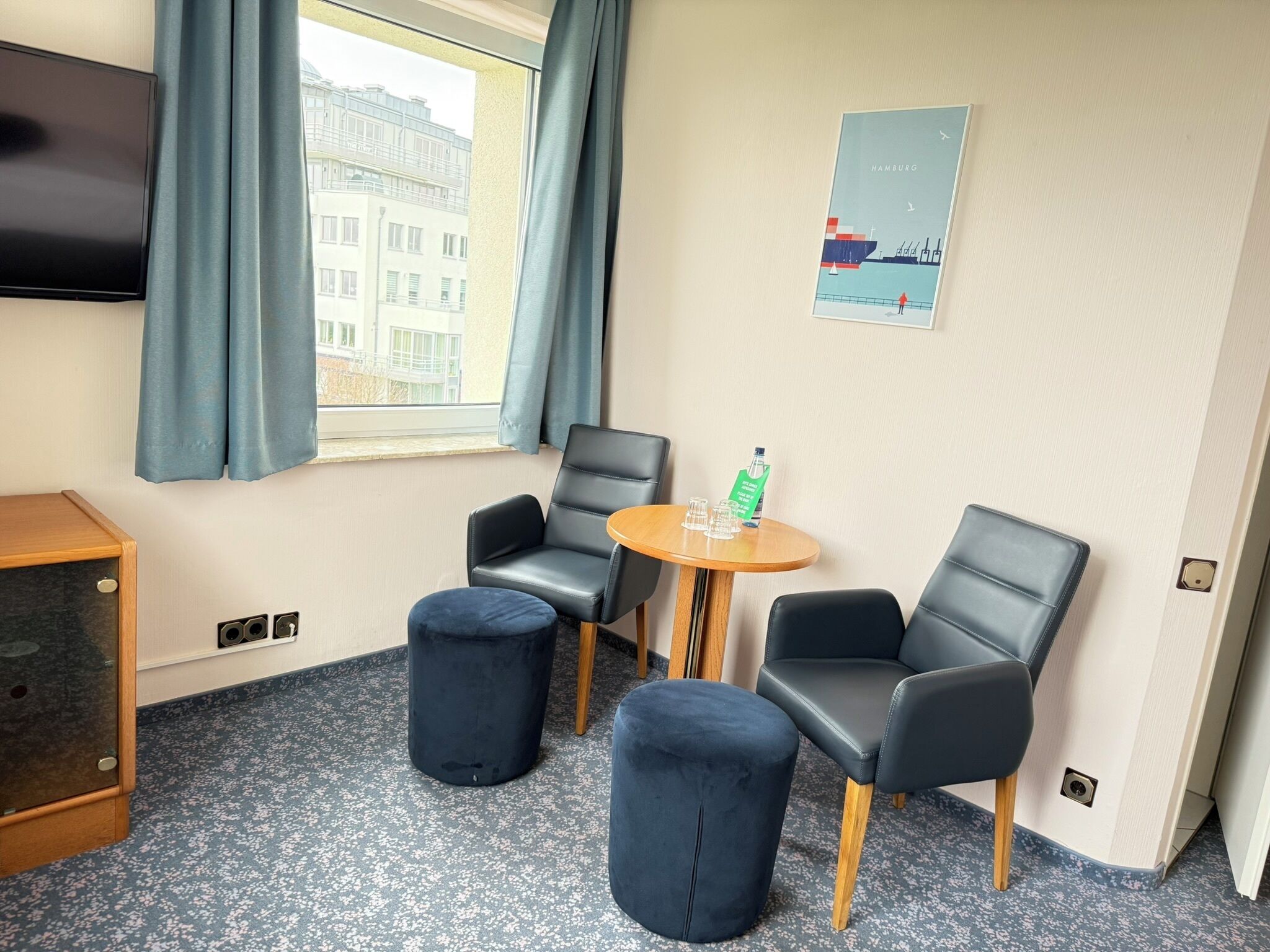 A hotel room with a TV, two black leather chairs, a small round table with glasses and a bottle, and a poster of Hamburg on the wall.