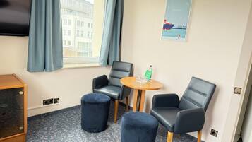 Een hotelkamer met een televisie, twee zwarte leren fauteuils, een klein rond tafeltje met glazen en een fles, en een poster van Hamburg aan de muur.