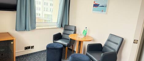 A hotel room with a TV, two black leather chairs, a small round table with glasses and a bottle, and a poster of Hamburg on the wall.