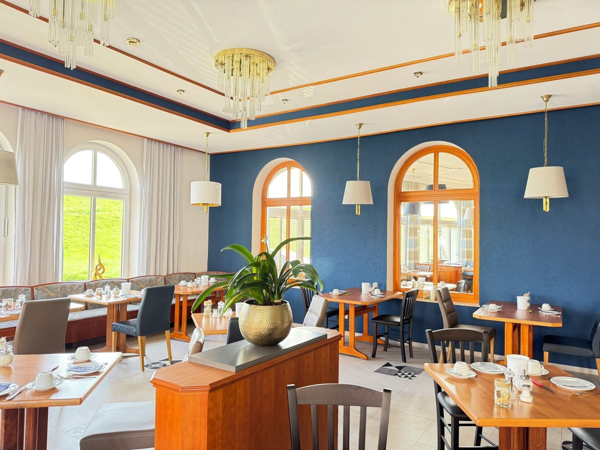 Zona de menjador d'hotel amb diverses taules i cadires, una taulell de recepció de fusta i grans finestres amb cortines.