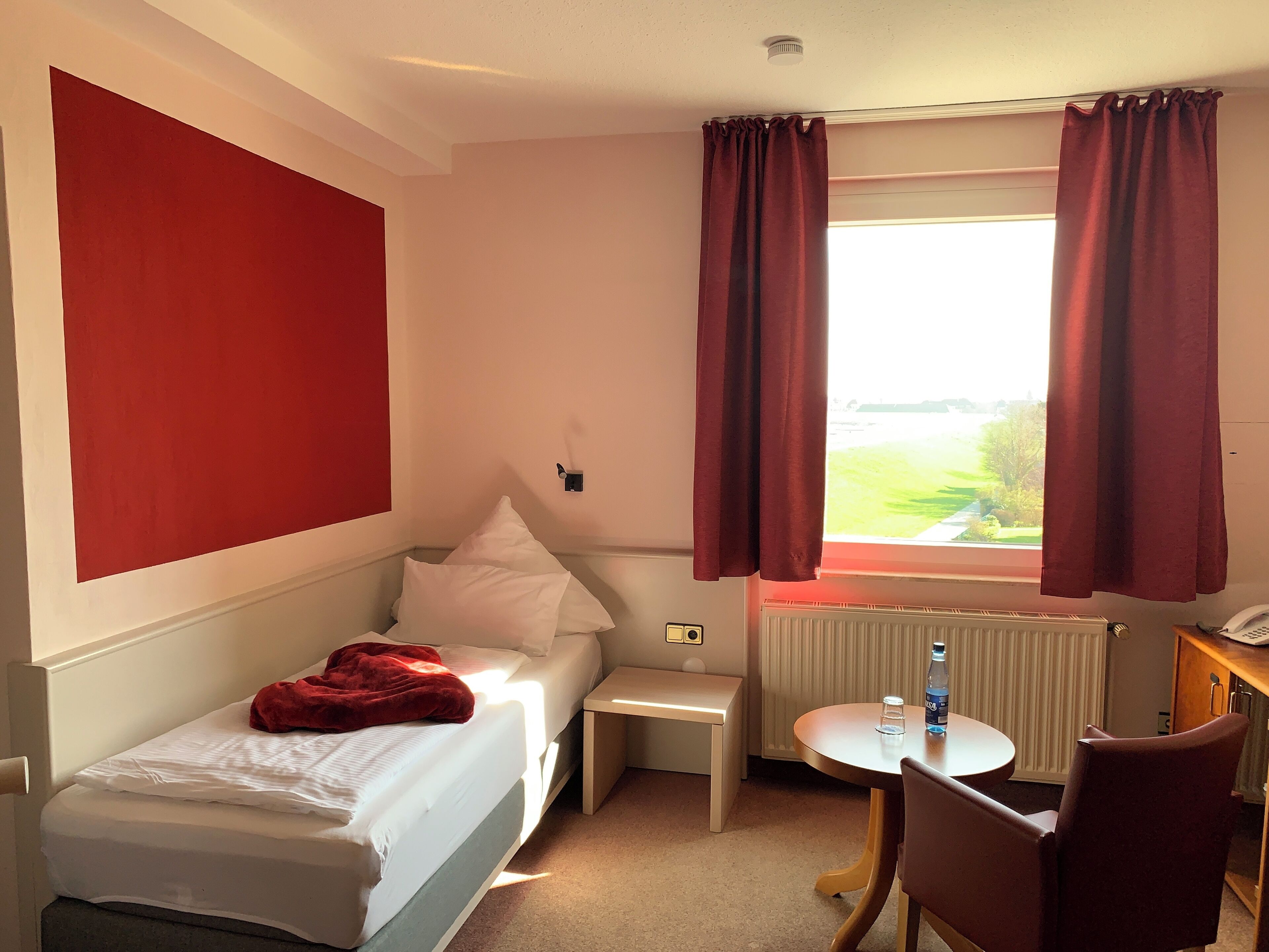 Une chambre d’hôtel avec un lit, une petite table, une chaise, une fenêtre avec des rideaux et une vue sur l’extérieur.