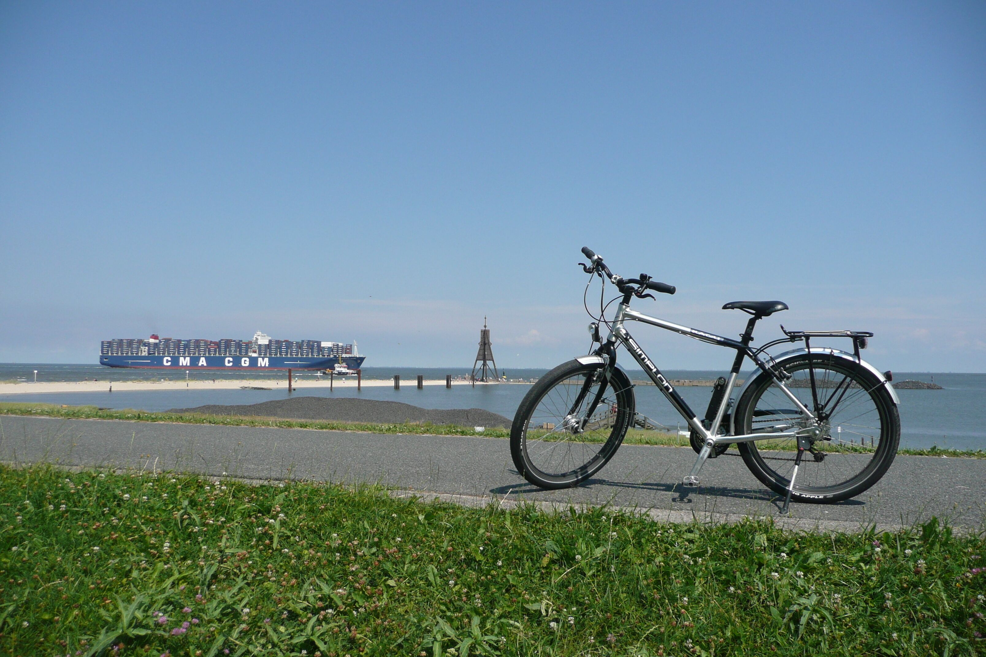 Ein Fahrrad steht auf einem gepflasterten Weg am Meer, im Hintergrund ein Frachtschiff.