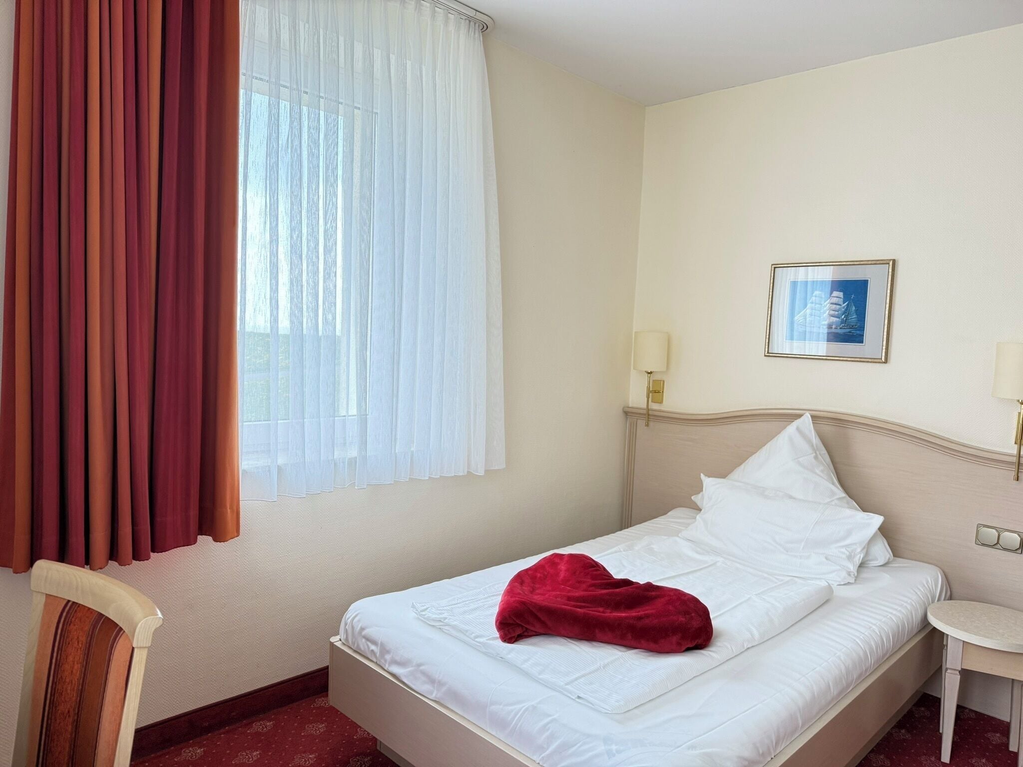 Hotellihuone, jossa on sänky, tuoli, pieni pöytä, ikkuna verhoilla ja seinällä oleva kehystetty kuva.