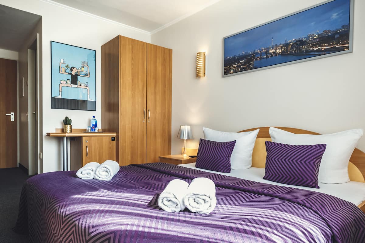 A hotel room with a bed, a desk, a nightstand, and a cityscape painting.