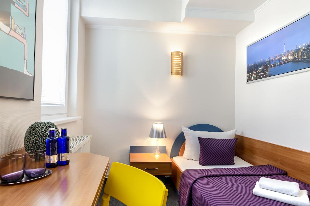 A hotel room with a bed, bedside table, lamp, and a cityscape painting.