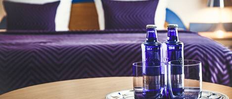 A hotel room with a bed, pillows, and a tray table with two bottles and glasses.