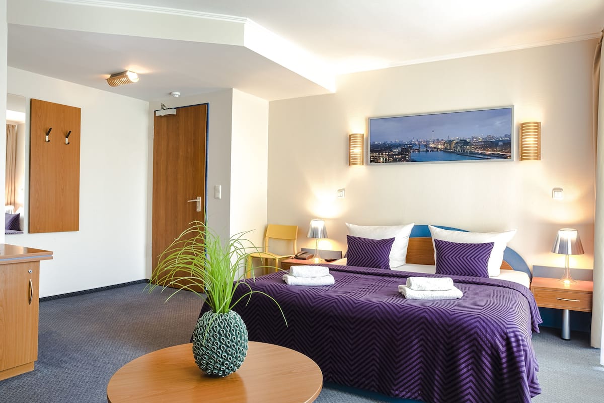 A hotel room with a bed, a round table with a pineapple centerpiece, a cityscape painting, and a wooden door.