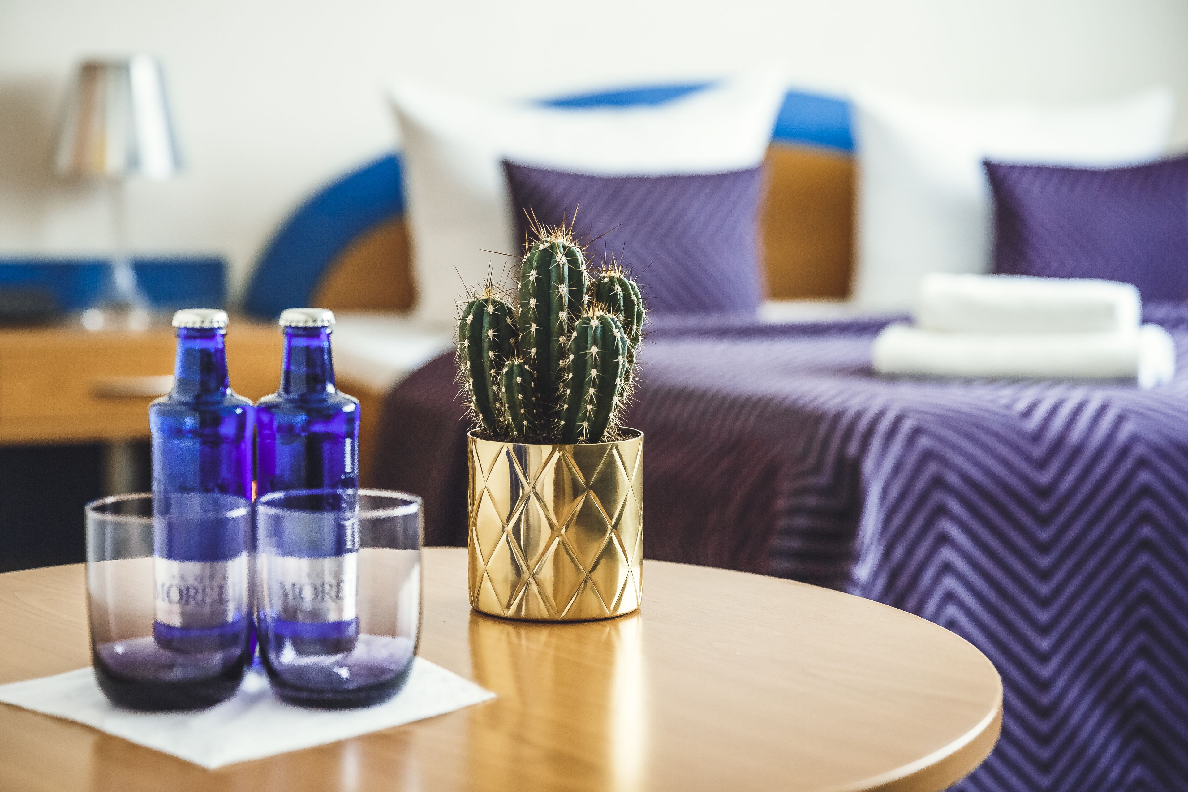 A bed with a purple and white bedspread, a gold decorative vase with a cactus, and two blue bottles on a table.