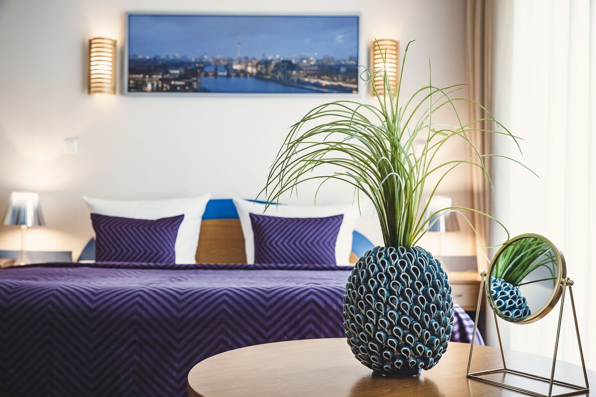 Hotellihuone, jossa on sänky, pyöreä peili, ananaksesta tehty koriste ja kaupunkimaisemaa esittävä taulu seinällä.