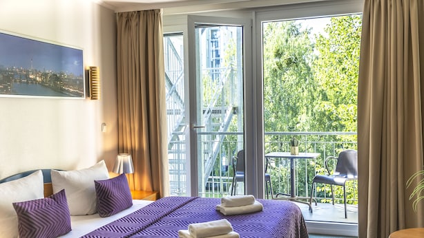 A hotel room with a bed, pillows, a nightstand, a window with curtains, a balcony with chairs, and a table. - Hotel Ambiente Berlin City (Berlin)