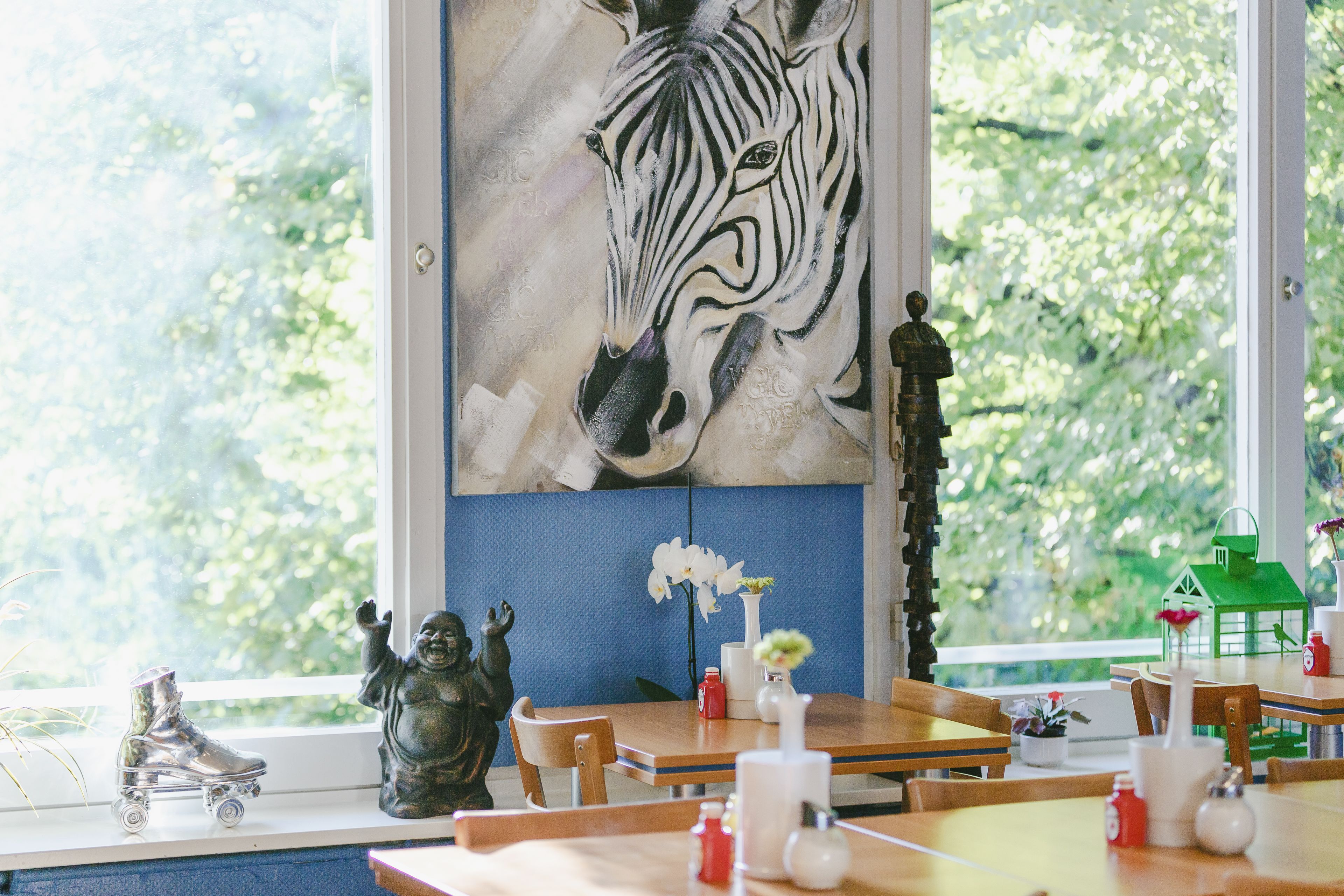 Ein Essbereich mit großem Zebra-Gemälde an der Wand, Holztischen, Stühlen und verschiedenen dekorativen Objekten, darunter ein kleiner Rollschuh, eine Figur und eine grüne Laterne.