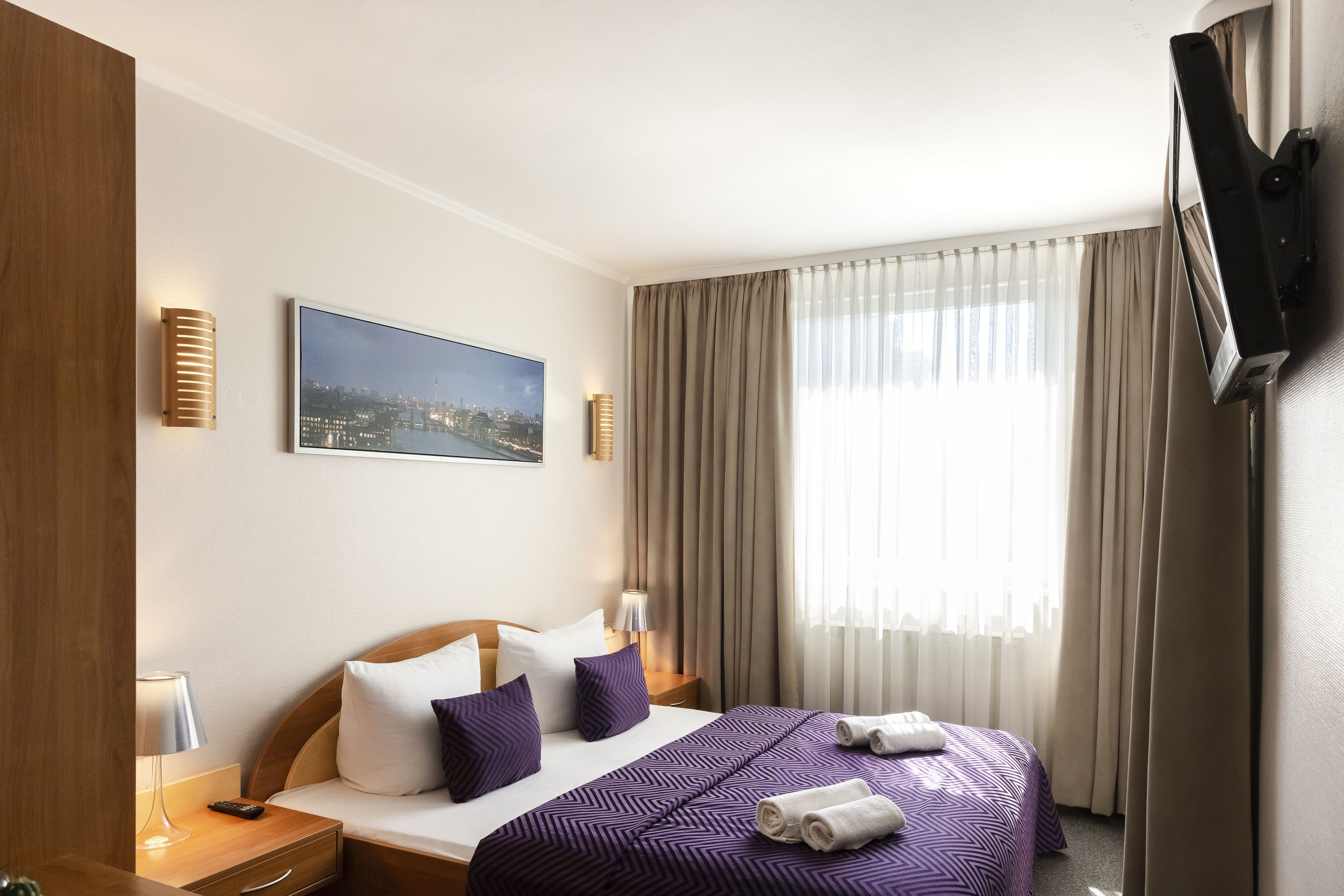 Una habitación de hotel con una cama, un televisor, un cuadro de paisaje urbano y una ventana con cortinas.