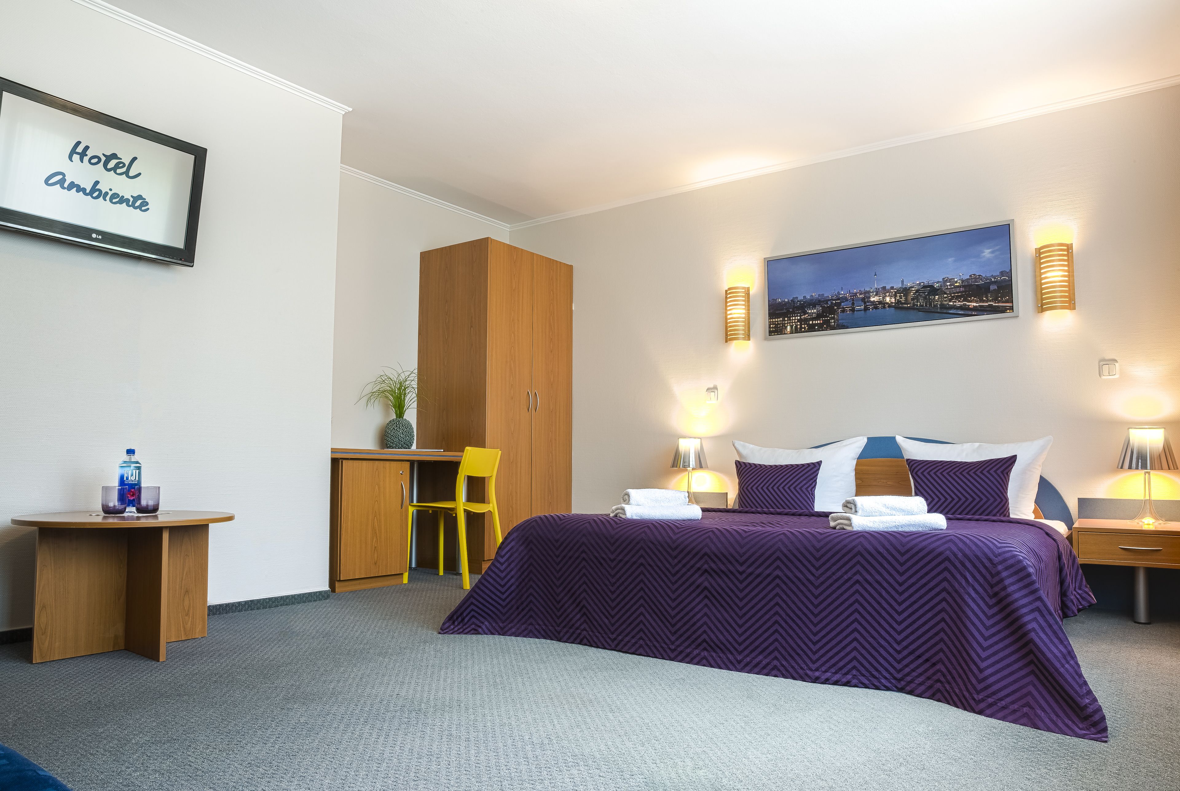 A hotel room with a bed, a desk, a chair, a wardrobe, and a nightstand with a lamp.
