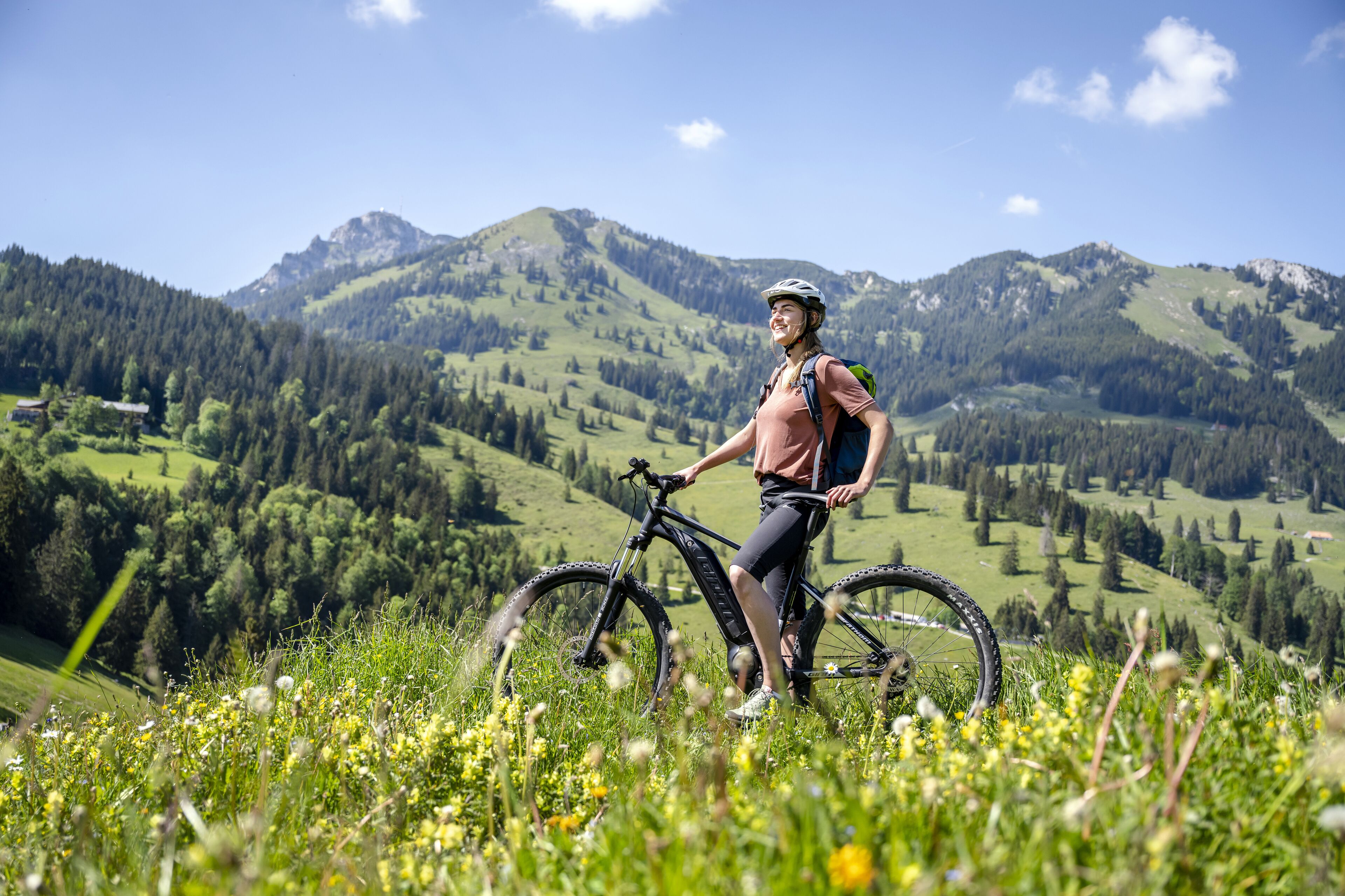 Eine Person steht neben einem Mountainbike auf einer grasbewachsen Anhöhe, im Hintergrund eine Berglandschaft.