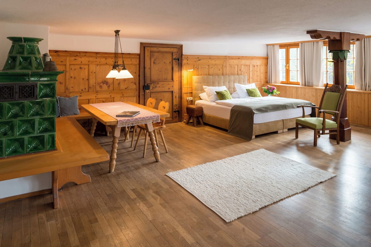 Een knus vertrek met een houten bed, een eettafel met stoelen, een groene kachel en een raam met gordijnen.