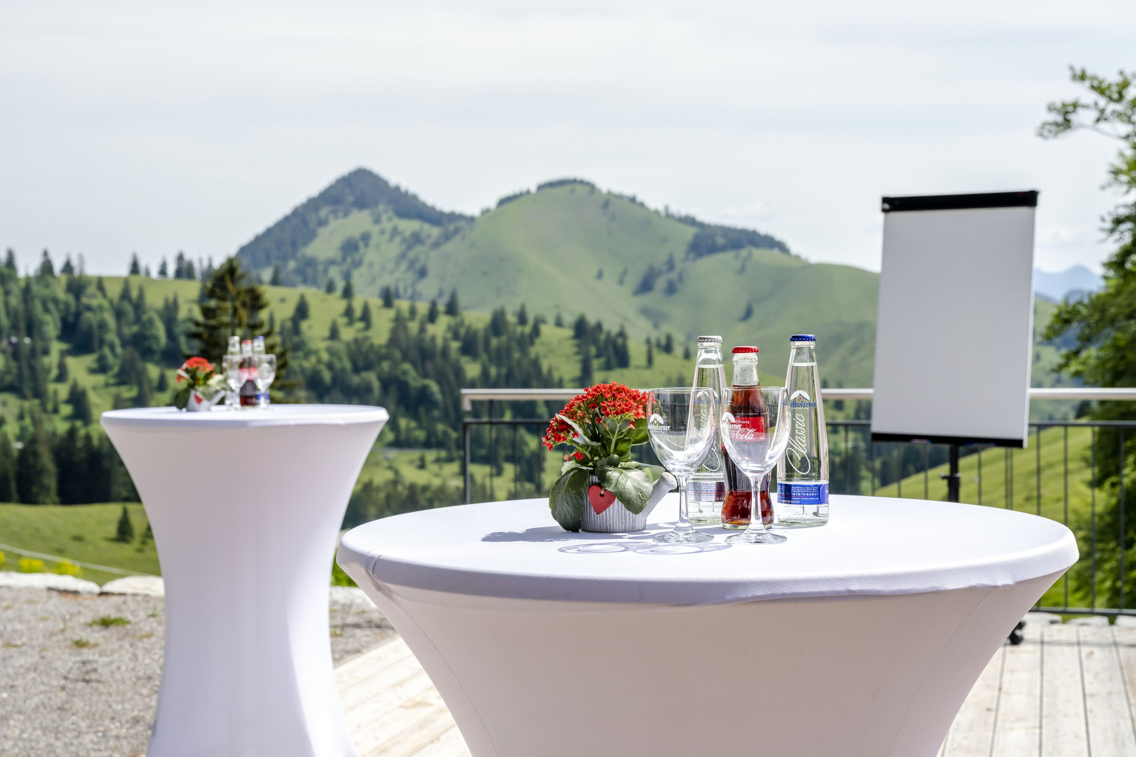 Two round tables with white tablecloths, each with a vase of red flowers, wine glasses, and bottles of water and wine. A projector screen is visible in the background.