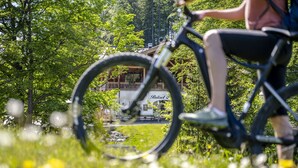 Una persona montando en bicicleta por un área con césped, árboles y un edificio al fondo.