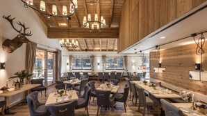 Un comedor amplio con vigas de madera, candelabros y una gran cabeza de ciervo montada en la pared. Las mesas están dispuestas con manteles blancos y velas.