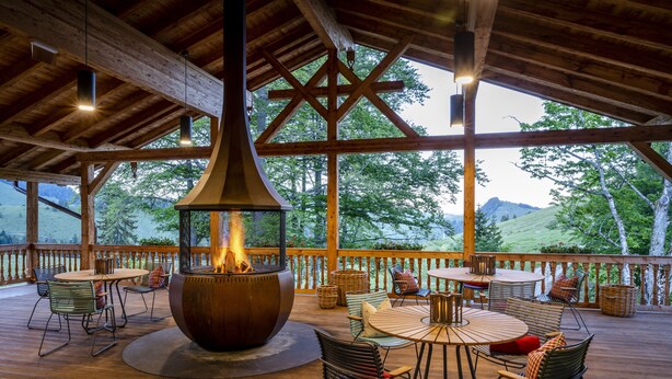 Una terraza de madera con mobiliario exterior, una chimenea en el centro y vistas del paisaje circundante.