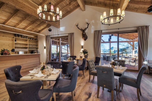 Un restaurant avec des plafonds en bois, des lustres et une tête de cerf accrochée au mur. Les tables sont dressées pour le dîner, avec des chaises.