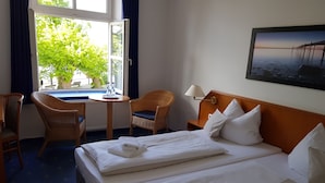 A hotel room with two beds, a desk, a chair, a lamp, and a window with a view of trees and a building. - Hotel Havenhaus (Bremen)