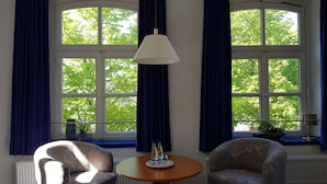 A room with two patterned armchairs, a round wooden table, and a floor lamp. There are large windows with curtains and a view of green trees. - Hotel Havenhaus (Bremen)