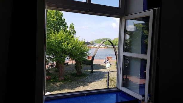 A room with a large window overlooking a riverside area with trees and a bridge. - Hotel Havenhaus (Bremen)