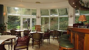 Un espace repas comprenant plusieurs tables et chaises, de grandes fenêtres donnant sur un paysage verdoyant, et un bar avec des tabourets.