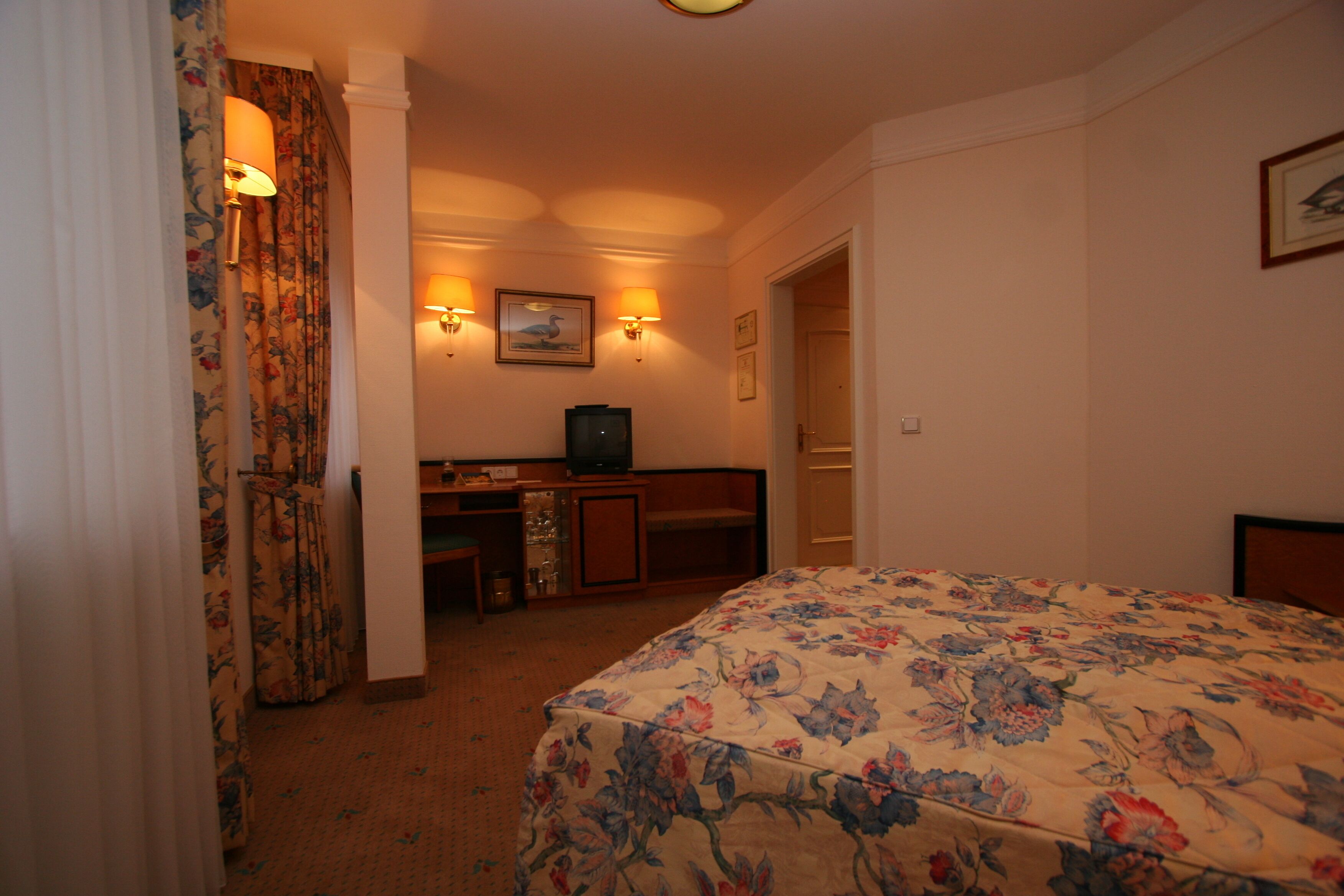 A hotel room with a floral bedspread, a desk with a television, and two wall-mounted lamps.
