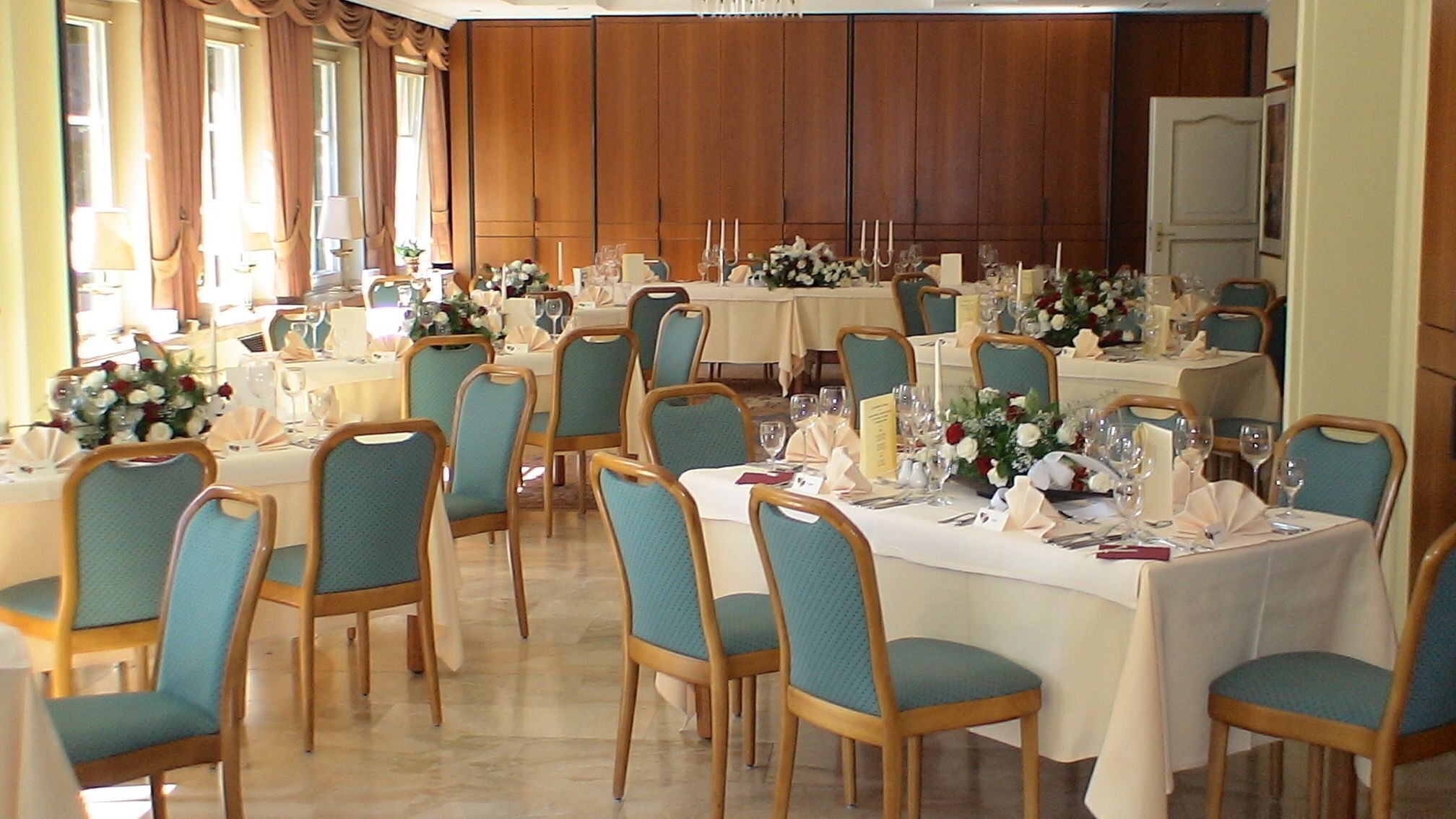 A formal dining room with multiple tables set for service, wooden chairs, and floral centerpieces.