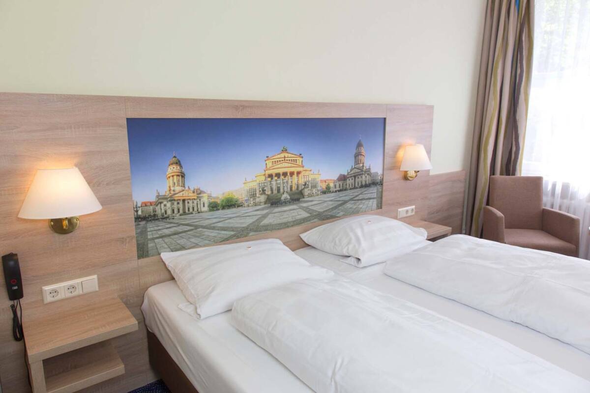 Quarto de hotel com duas camas, um grande mural na parede de um edifício histórico, abajures, uma cadeira e telefone ao lado da cama.