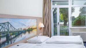Habitación de hotel con cama, un cuadro de un puente, una lámpara y vista a zonas verdes por la ventana.