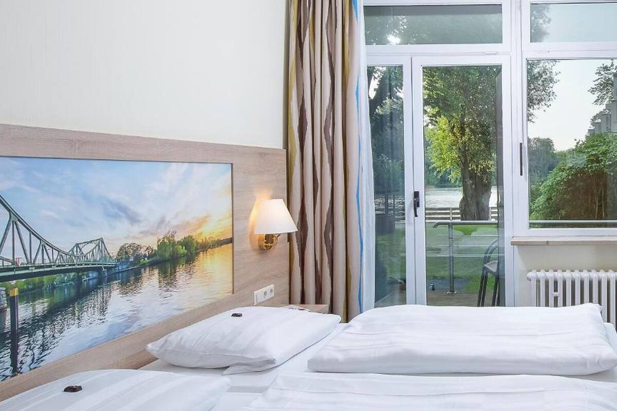 Quarto de hotel com uma cama, um quadro de uma ponte, uma luminária e vista para o verde através de uma janela.